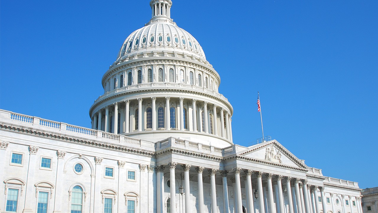 The U.S. Capitol building in Washington, D.C., with its prominent white dome and columns, set against a clear blue sky and an American flag flying.