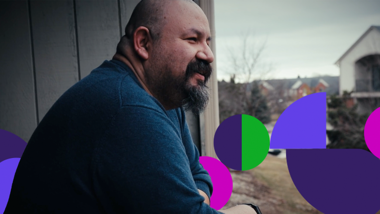 Man with a beard and mustache wearing a blue shirt leans on a balcony railing, smiling while looking into the distance, with colorful graphic shapes overlaying the foreground.