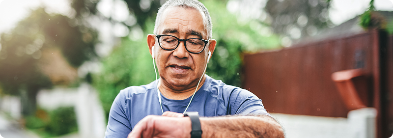 An older man wearing glasses and earphones checks his smartwatch while out for a walk or jog in a suburban neighborhood.