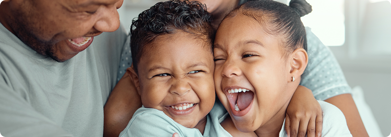 A father smiling joyfully as he hugs his two young children, who are laughing and cuddling close together.