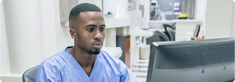 Image Alt Text:
A man wearing blue scrubs in a hospital setting looks at a black computer monitor with a neutral face.