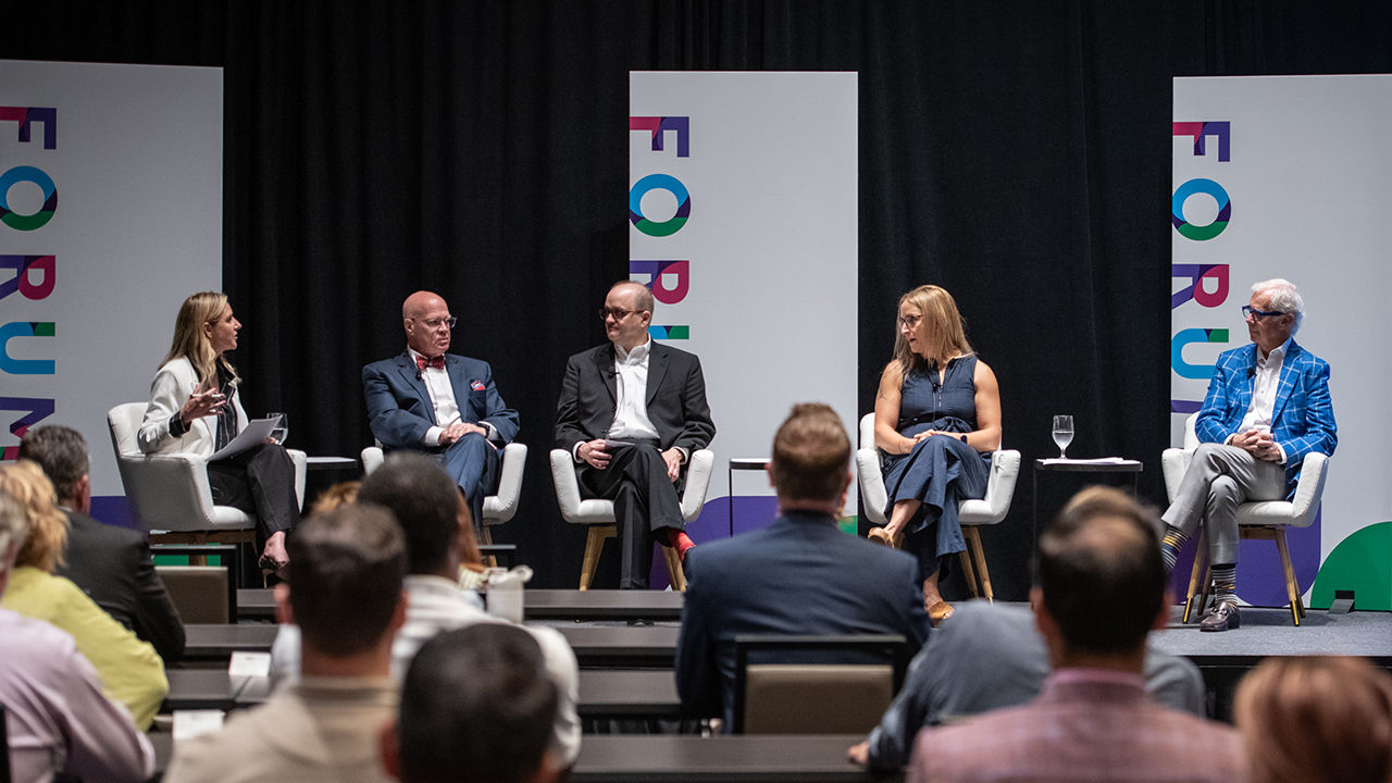 Five professionally dressed panelists sit on stage in white chairs having a discussion during Forum 2025. Three panels behind them display the event name in bold, colorful letters on white background.