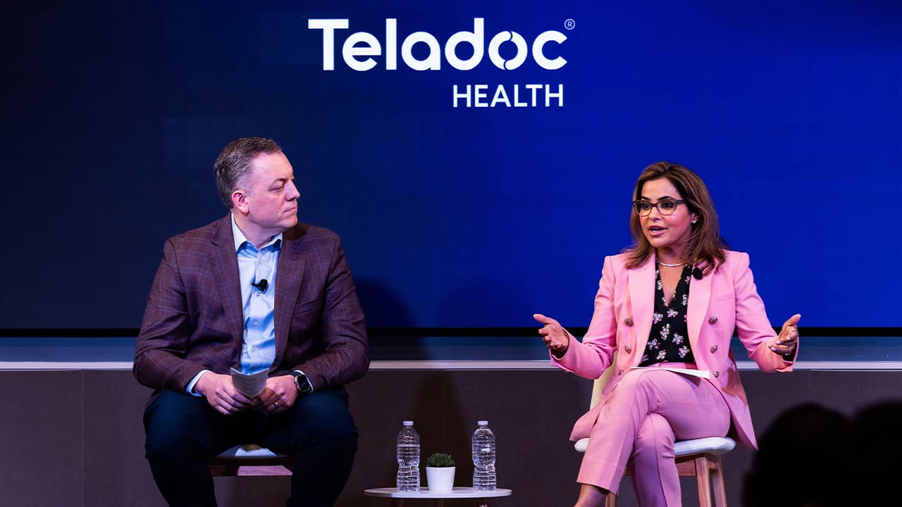 Two speakers seated on a stage during a Teladoc Health event, participating in a panel discussion