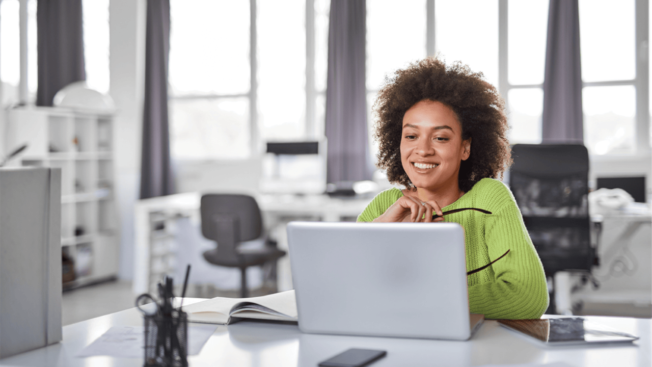 A cheerful young woman with curly hair sitting at a desk, working on a laptop