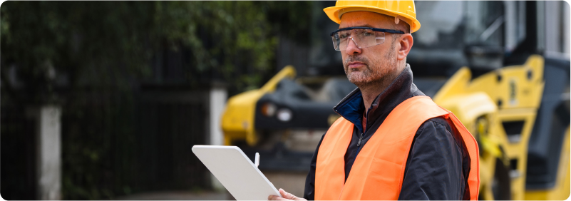 Construction worker in safety gear holding clipboard near road roller.