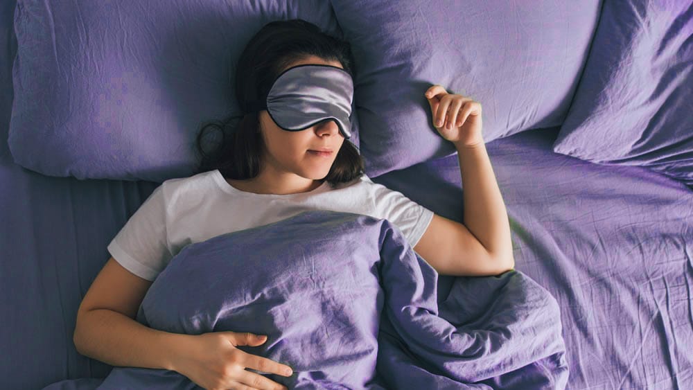 Woman sleeping in bed wearing an eye mask and covered with a blanket
