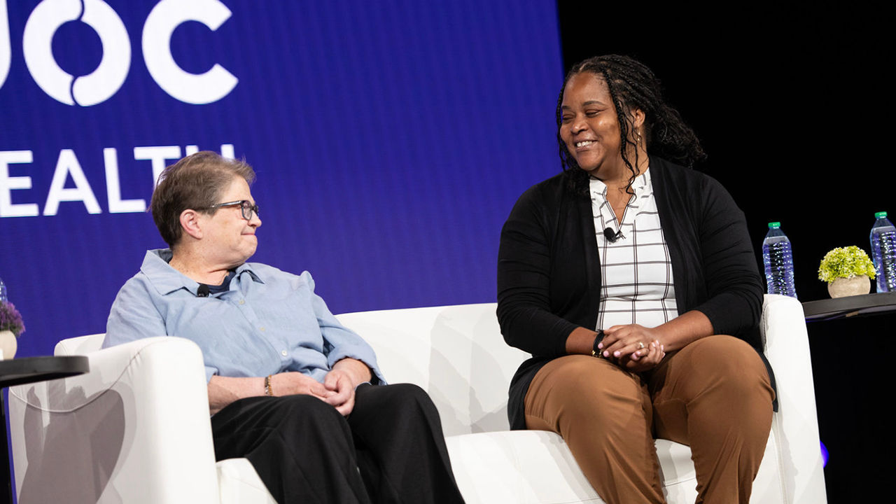 Pat and Keisha sitting on a white couch on a stage looking at each other with a large white Teladoc Health logo on a dark purple panel in the background.