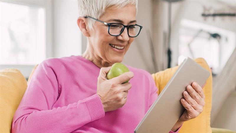 Woman with short gray hair wearing glasses and a pink sweater, holding a green apple and looking at a tablet while sitting on a couch.