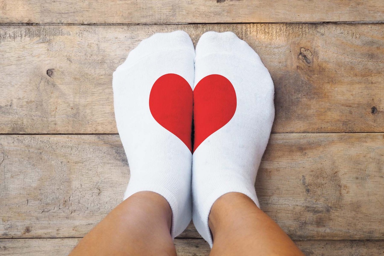 Pair of feet wearing white socks with a red heart design that forms when both feet are placed together, resting on a wooden floor.