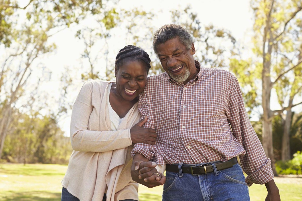 Happy older couple walking arm-in-arm outdoors in a park, laughing and enjoying each other's company with trees and sunlight in the background.
