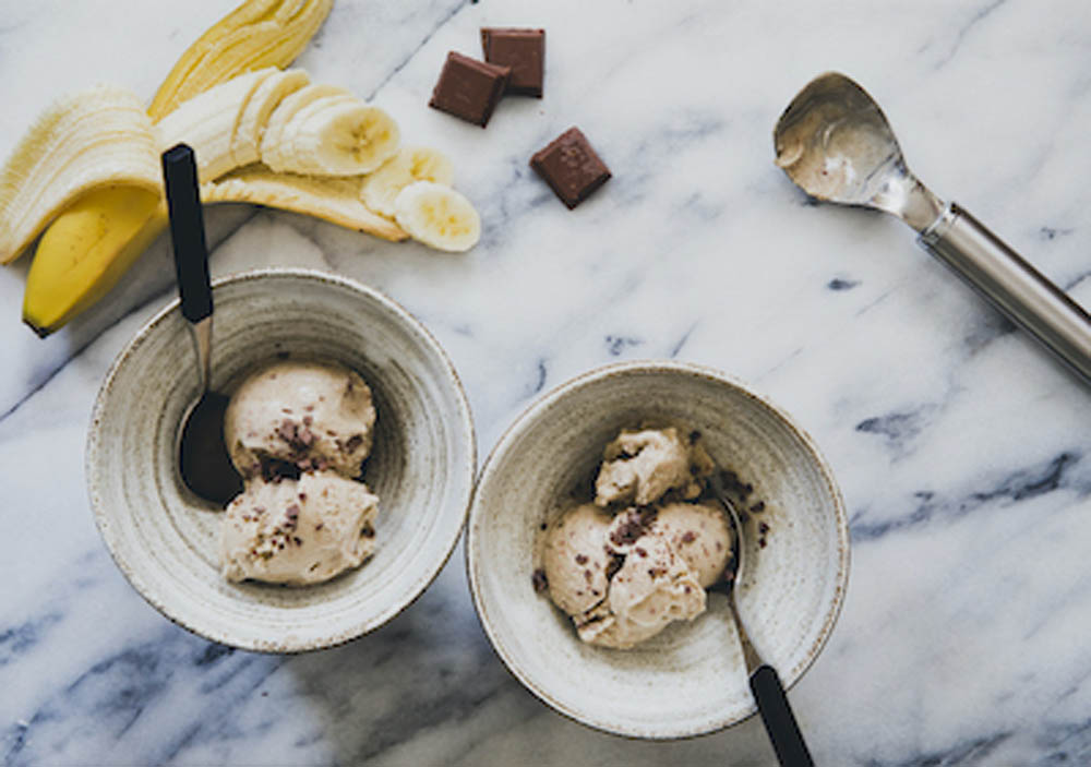 Two bowls of ice cream with chocolate chips, surrounded by sliced bananas, chocolate pieces, and an ice cream scooper on a marble surface.