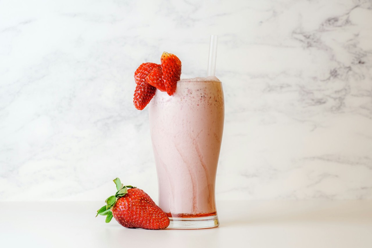 A tall glass filled with a creamy strawberry smoothie, topped with fresh strawberries, and a straw, accompanied by a couple of whole strawberries on the table, against a light marble background.