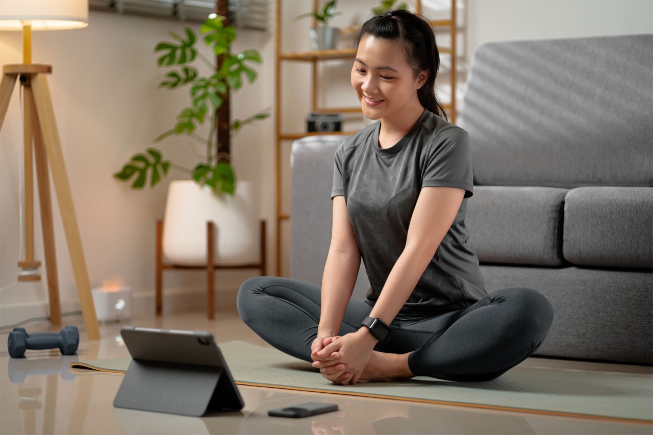 Smiling woman sitting cross-legged on a yoga mat, watching a tablet during an at-home workout session, with a cozy living room setting featuring a gray couch, indoor plants, and a floor lamp.