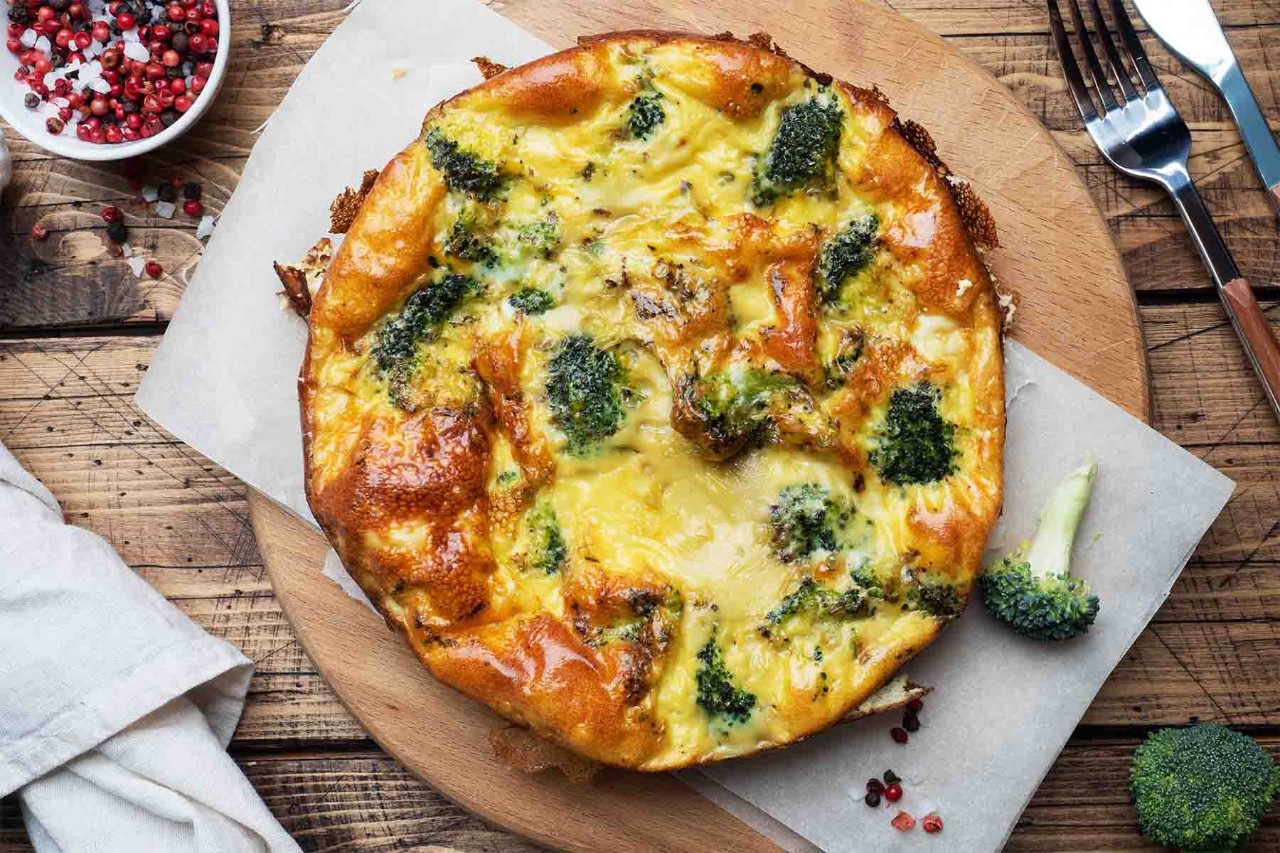 Golden-brown broccoli and cheese quiche with a flaky crust, served on parchment paper atop a wooden cutting board, with fresh broccoli florets and peppercorns scattered around.
