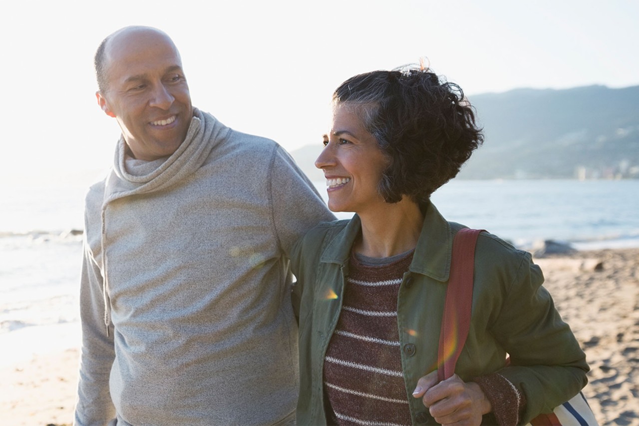Smiling couple walking arm in arm along a beach with mountains in the background.