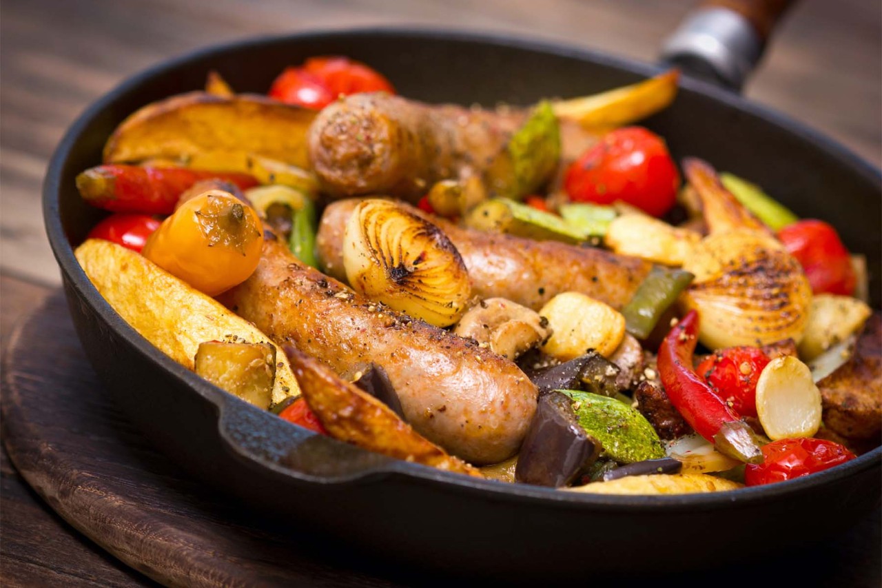 A cast-iron skillet filled with roasted sausages, cherry tomatoes, and assorted vegetables, including peppers and zucchini, on a wooden surface.