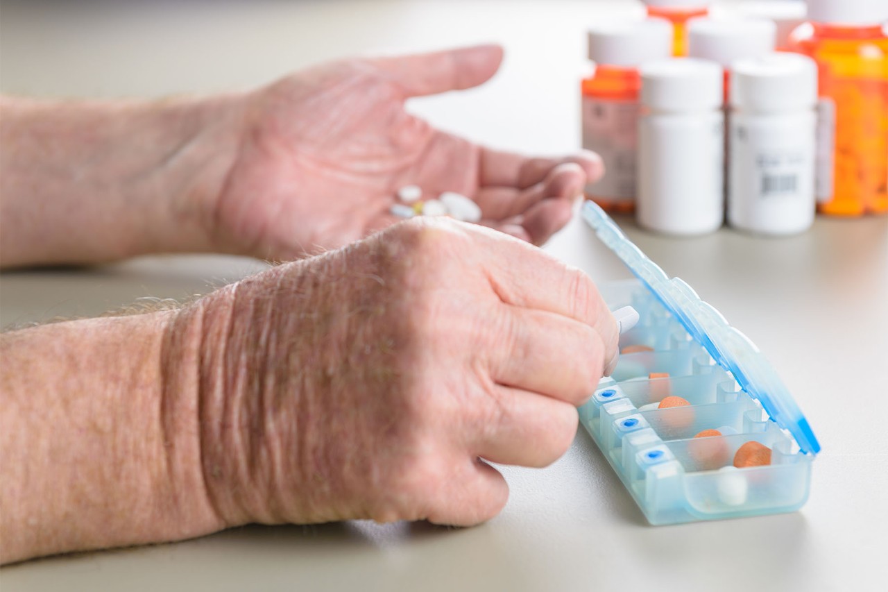 Close-up of an elderly person's hands placing pills into a weekly pill organizer, with prescription medication bottles in the background.