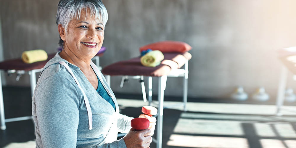 A smiling older woman with short gray hair, wearing a gray hoodie, holding red dumbbells in a fitness setting, with exercise mats and towels visible in the background.
