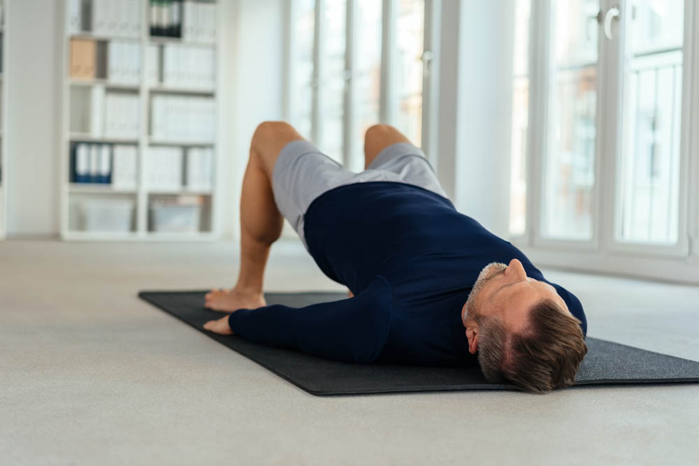Man lying on a yoga mat indoors, performing a glute bridge exercise with knees bent and hips lifted, in a bright room with large windows and shelves filled with binders.