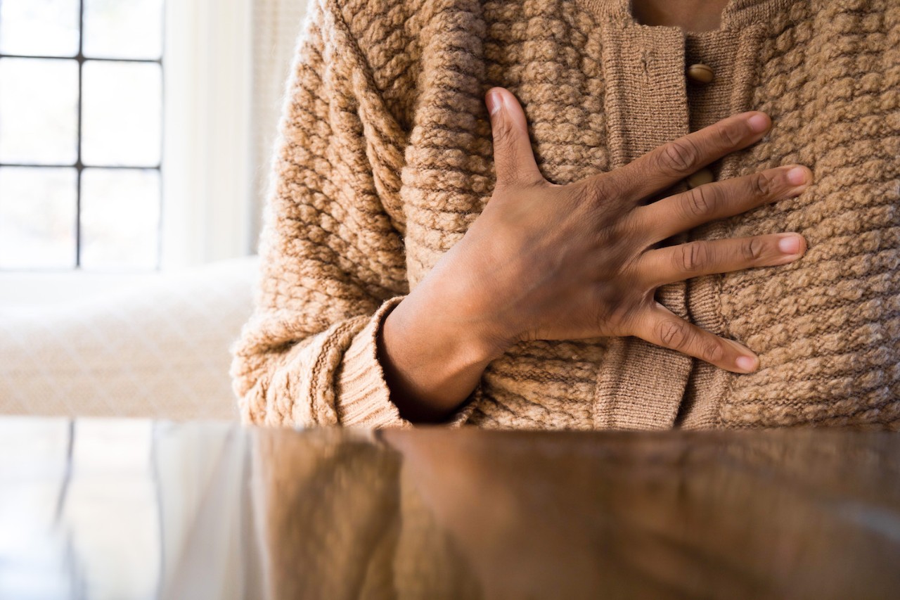 Person wearing a textured brown cardigan holding their hand over their chest while seated at a table with a window in the background.