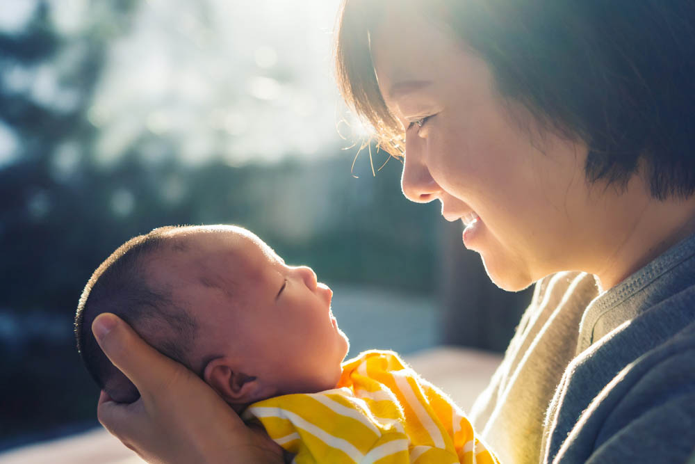  A close-up of a smiling woman holding a newborn baby, both gazing at each other warmly in a softly lit setting.