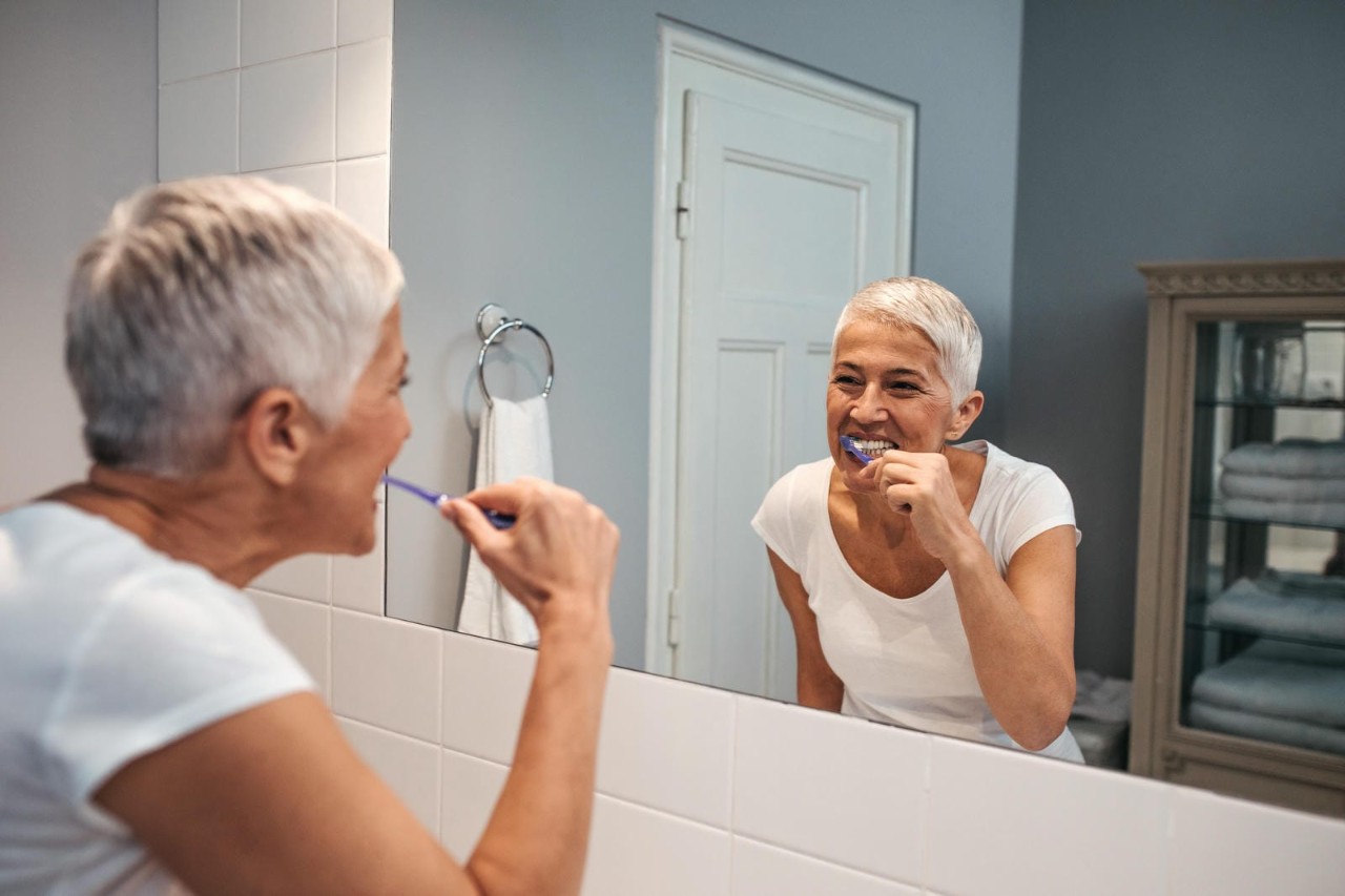 A woman with short, gray hair is brushing her teeth in front of a bathroom mirror, smiling at her reflection. The bathroom has a clean and modern appearance.