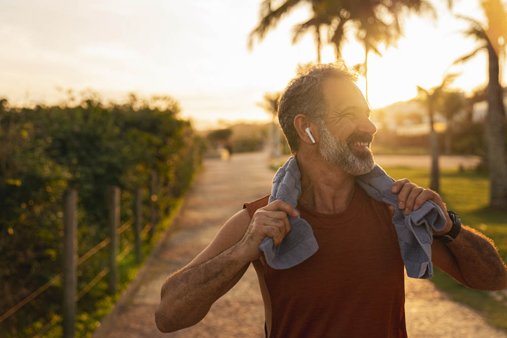 A motivated senior athlete, standing with a smartphone in his hand and a towel around his neck. He catches his breath after running and rests