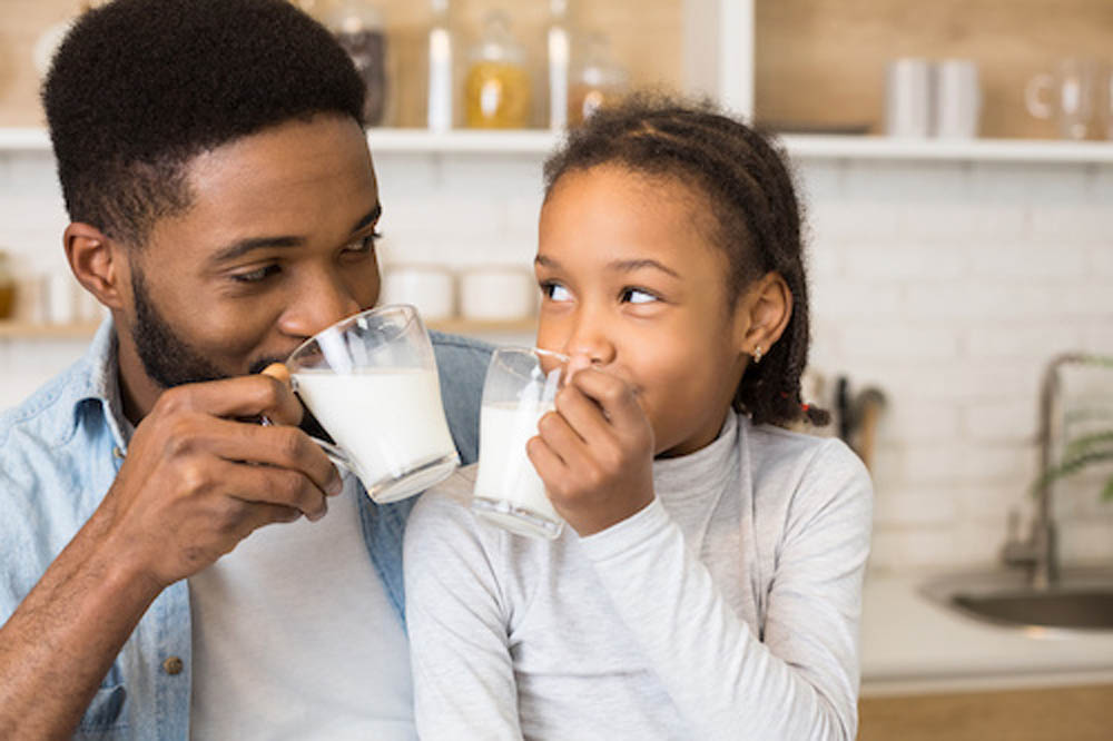 A father and daughter are smiling at each other while enjoying glasses of milk in a cozy kitchen.