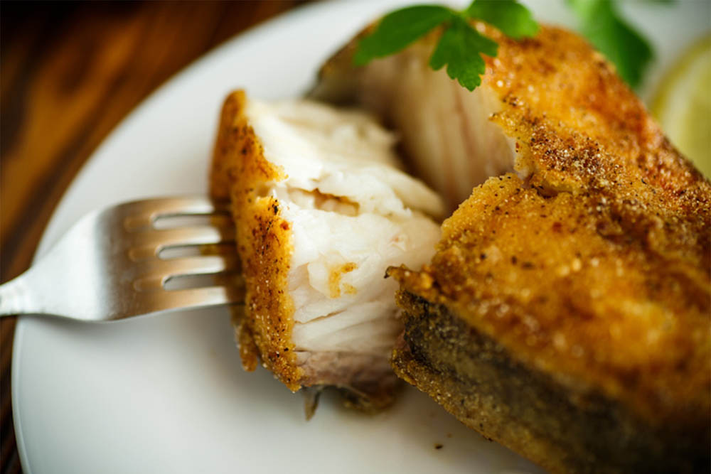 A close-up of a piece of golden-brown fried fish, with a fork piercing into it, revealing the tender white flesh inside. A sprig of fresh parsley and a slice of lemon are in the background.