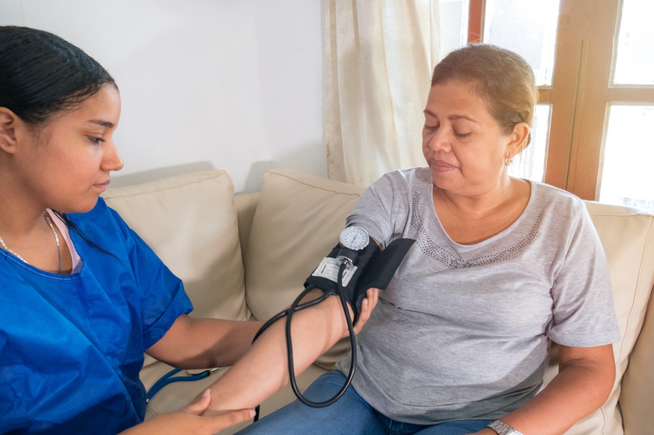 A healthcare worker measuring a womans blood pressure on a sofa in a well-lit room.