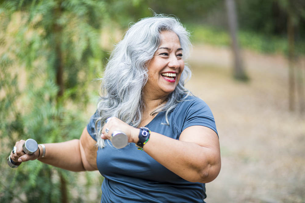 Smiling woman with gray hair exercises outdoors using small dumbbells, wearing a blue shirt and watch, surrounded by trees.