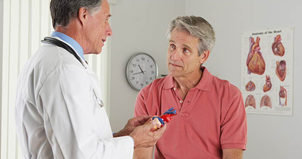 Doctor explaining heart model to a patient in an office with anatomical heart poster and clock in the background.