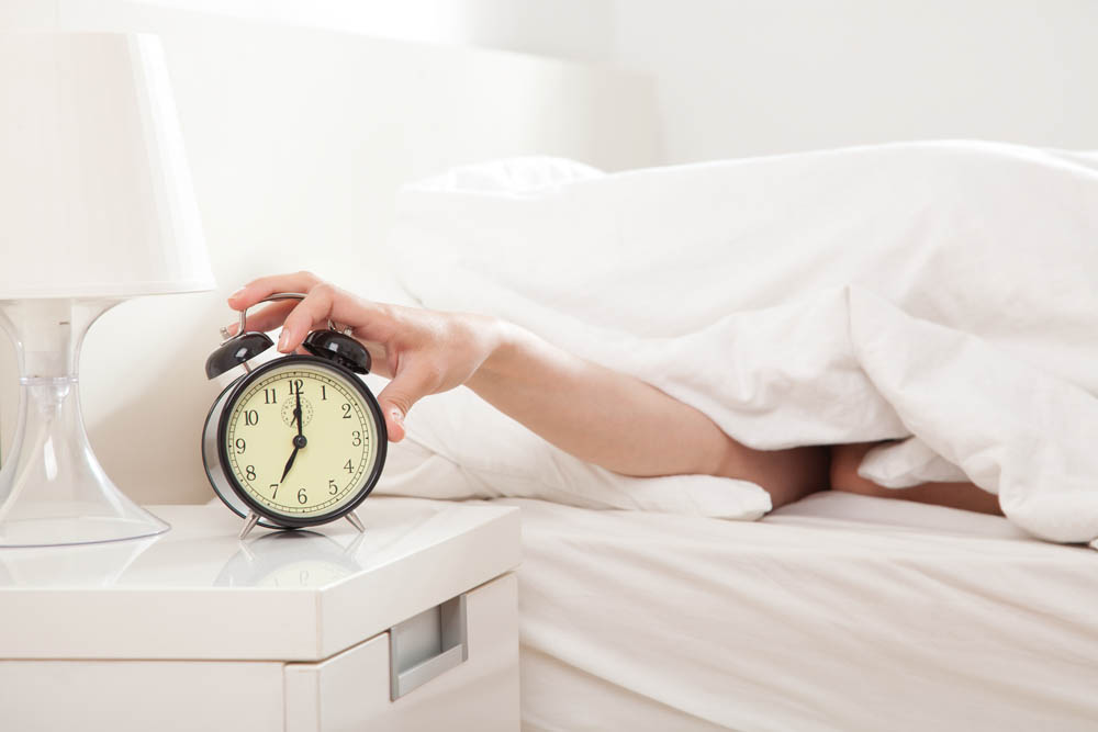 A hand reaching out from under a white duvet to turn off an alarm clock on a bedside table next to a white lamp. The time on the clock is 7:00.
