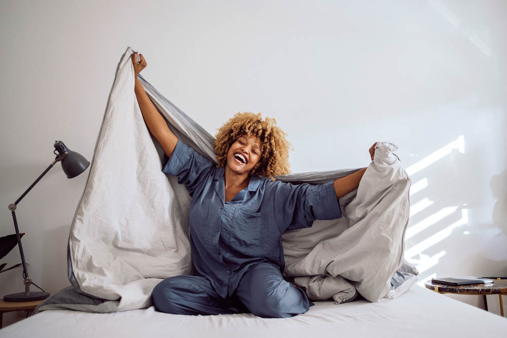 Woman in her pajamas smiling and sitting on the bed while playing with bed covers. She is holding them on her back like a cape.