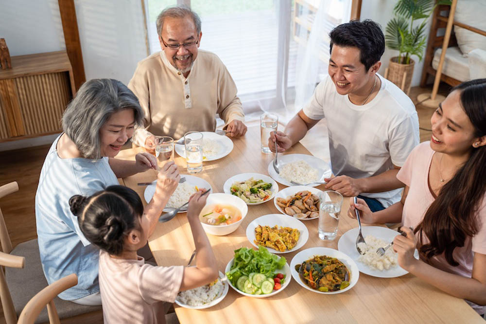 A family of six enjoying a meal together at a dining table with rice, vegetables, meat, and fruit.