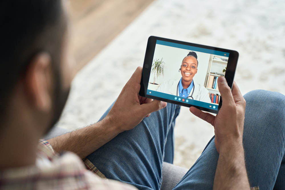 A man holding a tablet showing a video call with a doctor, sitting cross-legged on a couch.
