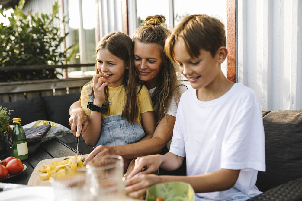 A woman and two children sit at an outdoor table, preparing food and smiling together.