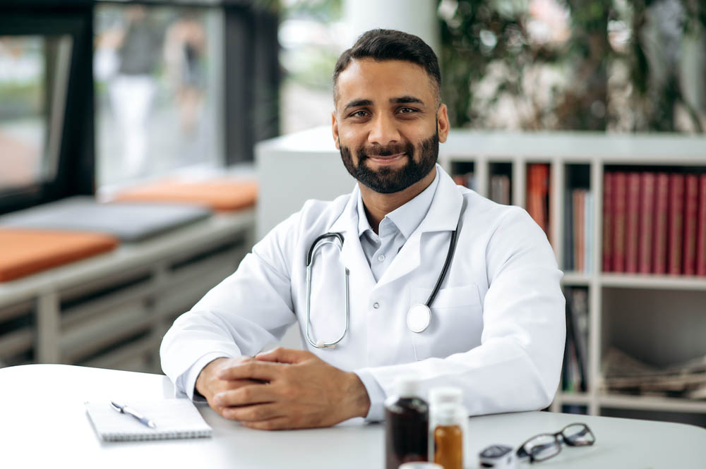 A doctor wearing a white coat and stethoscope sitting at a table in an office, smiling. Shelves with books and a window are in the background.