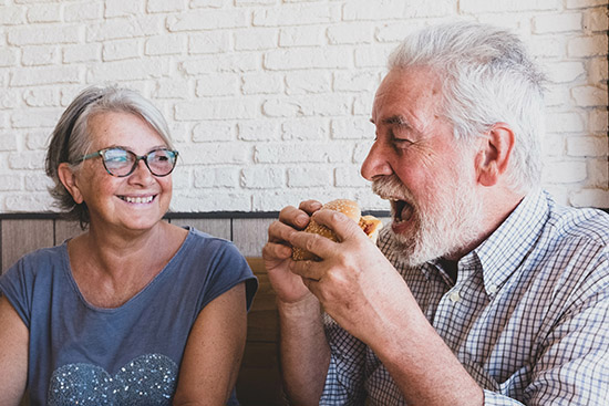Older man eating a sandwich, sitting next to a smiling woman with glasses, against a white brick wall.