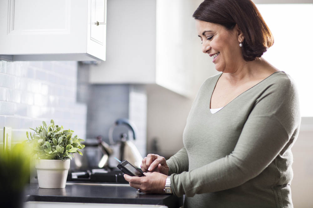 A smiling woman in a kitchen using a glucose meter to check her blood sugar levels.