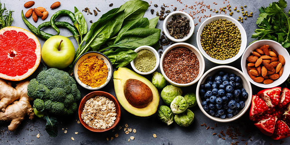 A spread of fresh fruits, vegetables, seeds, and nuts on a gray surface, including grapefruit, avocado, broccoli, blueberries, and almonds.