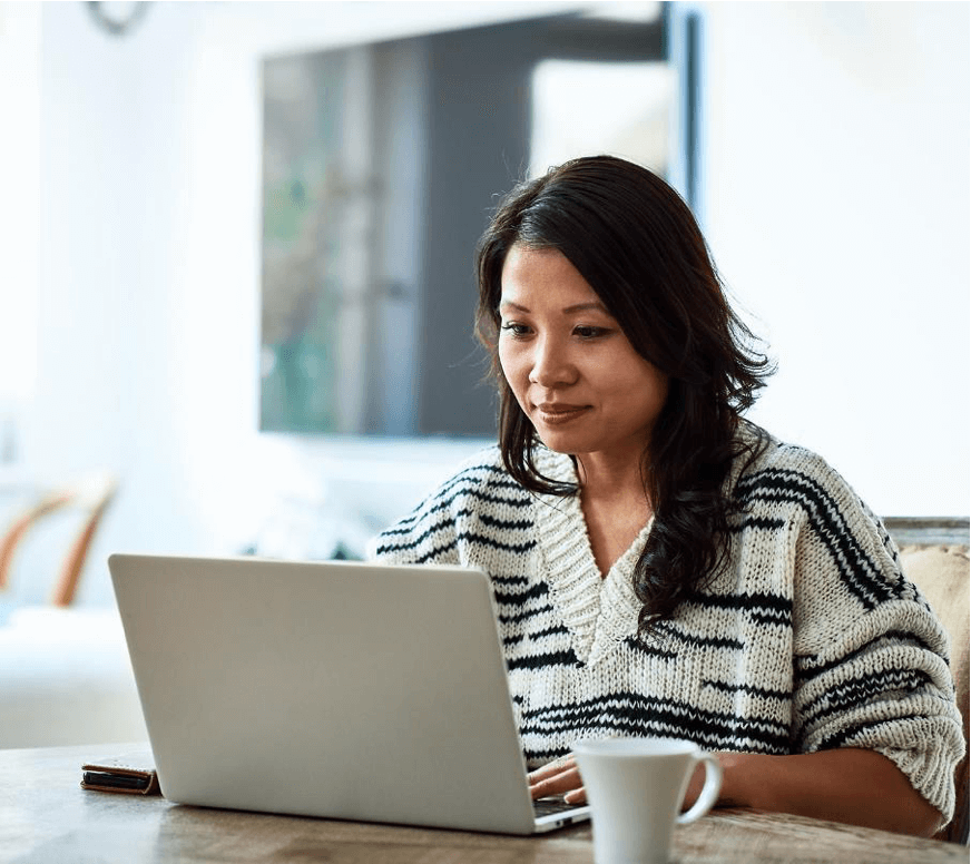 Woman in a striped sweater sitting at a table using a laptop, with a white mug in the foreground. Background shows a blurred television.