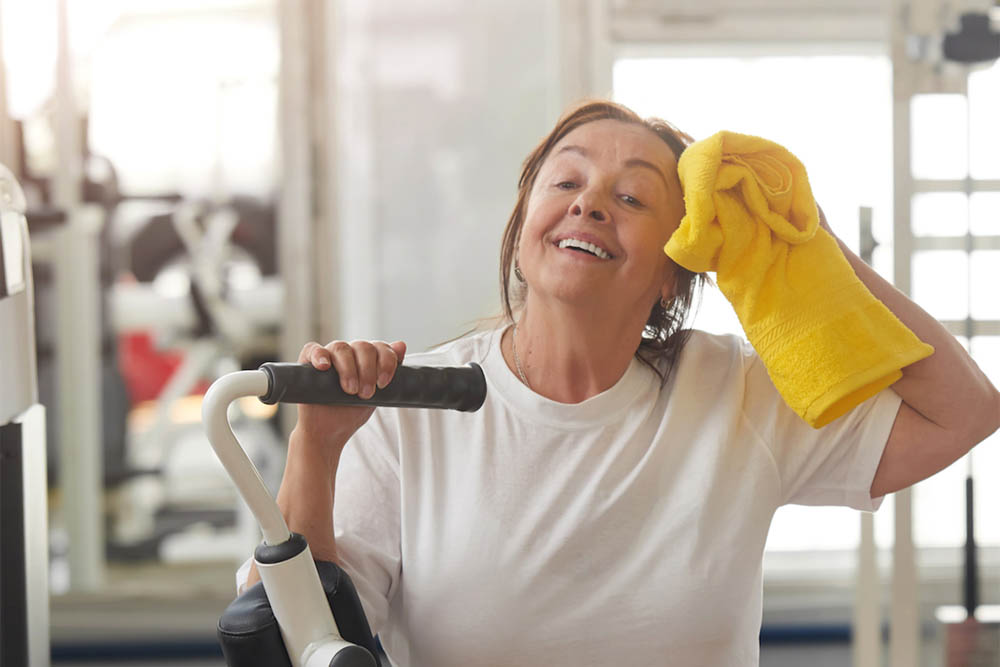 Mujer mayor sonriendo mientras se seca el sudor con una toalla amarilla en el gimnasio, apoyada sobre una máquina de ejercicio