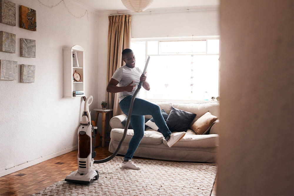  A person is joyfully vacuuming in a living room, dancing while holding the vacuum cleaner, with a cozy sofa and warm sunlight in the background.