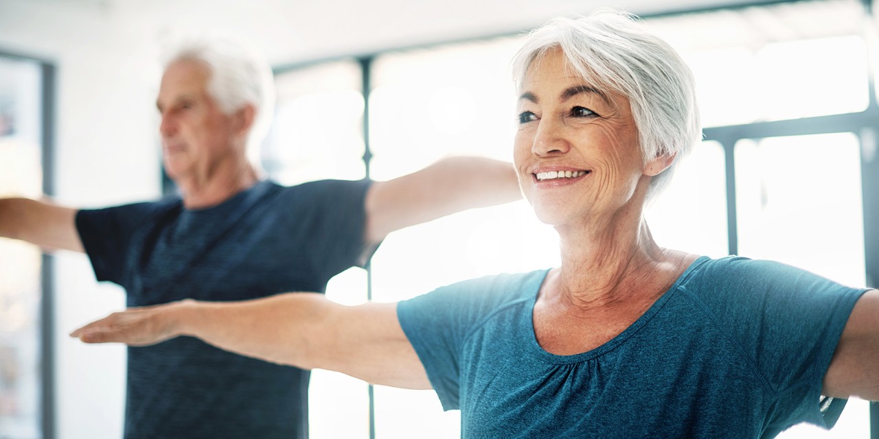 Two senior adults exercising indoors with outstretched arms, both wearing casual workout attire. They appear focused and content.