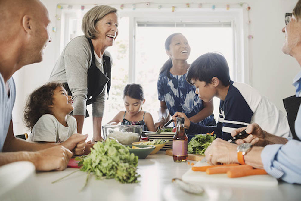 A diverse group of people, including adults and children, gather around a table with fresh vegetables, smiling and preparing food together.