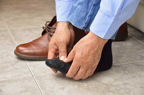 Person adjusting black sock on their foot, with brown leather shoe on tiled floor. Wearing blue jeans and a light blue shirt.