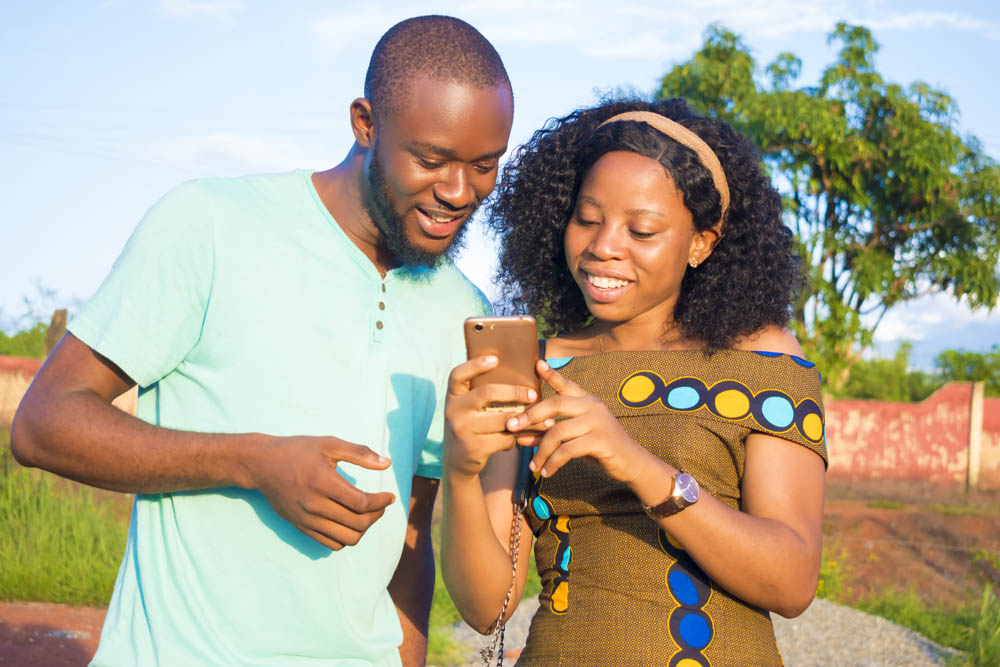 Two people smiling while looking at a smartphone outdoors, with trees and a wall in the background.