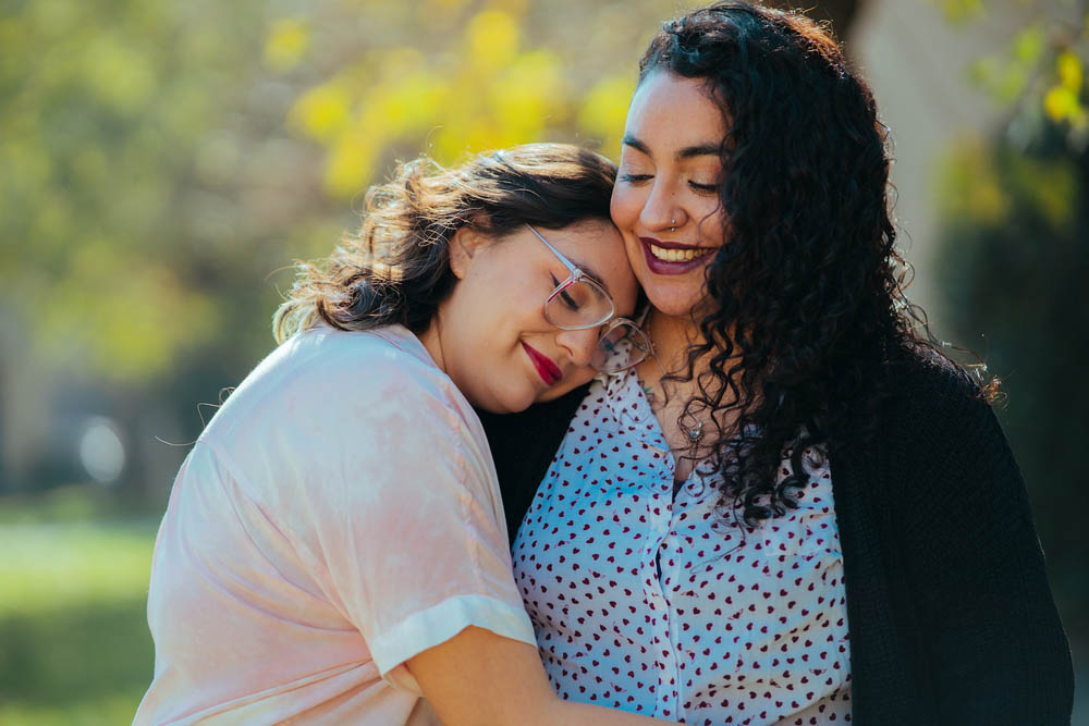 Two people embrace outdoors, one resting their head on the others chest, both smiling.