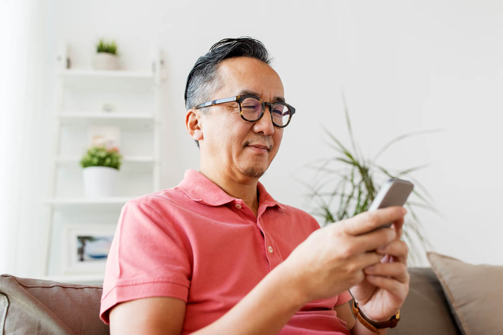 technology, people and communication concept - asian man with smartphone sitting on sofa at home